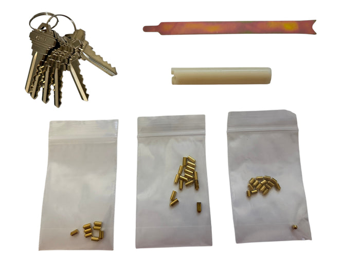 A set of lock pick tools and components laid out on a white background, including a keyring with several keys, a flat metal tool, a beige cylindrical piece, and three small clear bags containing small brass-colored hardware pieces.